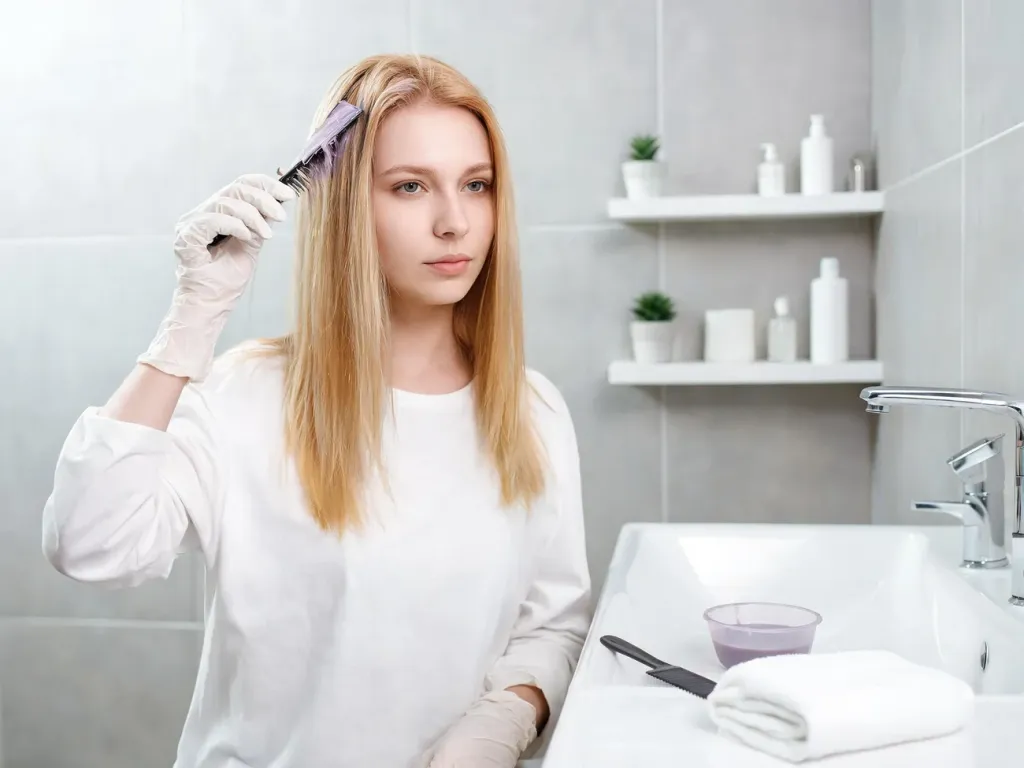 Kobieta w nowoczesnej łazience nakłada pędzelkiem toner na blond włosy, obok przygotowane akcesoria do koloryzacji.